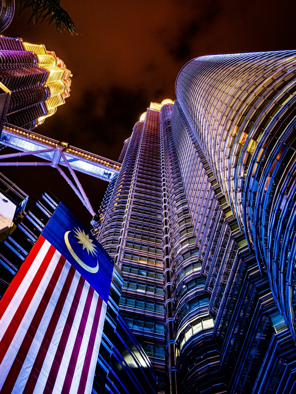 Dramatic view of Petronas Twin Towers illuminated at night with the Malaysian flag.